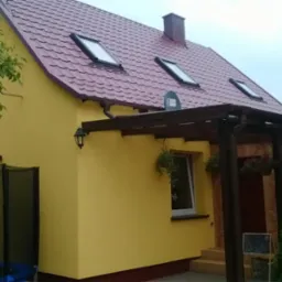 Żółty dom z bordowym dachem z dachówkami, trzema oknami dachowymi i drewnianą pergolą nad wejściem. Widoczny fragment ogrodu z trampoliną.