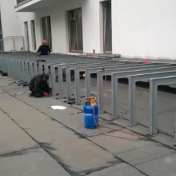 Prace dekarskie na dachu: spawanie metalowych konstrukcji pod platformę. Widoczny palnik, butla gazowa i dekarz w trakcie pracy.