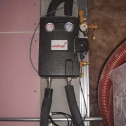 Czarny moduł Windhager z manometrami i rurami w piwnicy, widoczne elementy instalacji grzewczej i rury wentylacyjne.