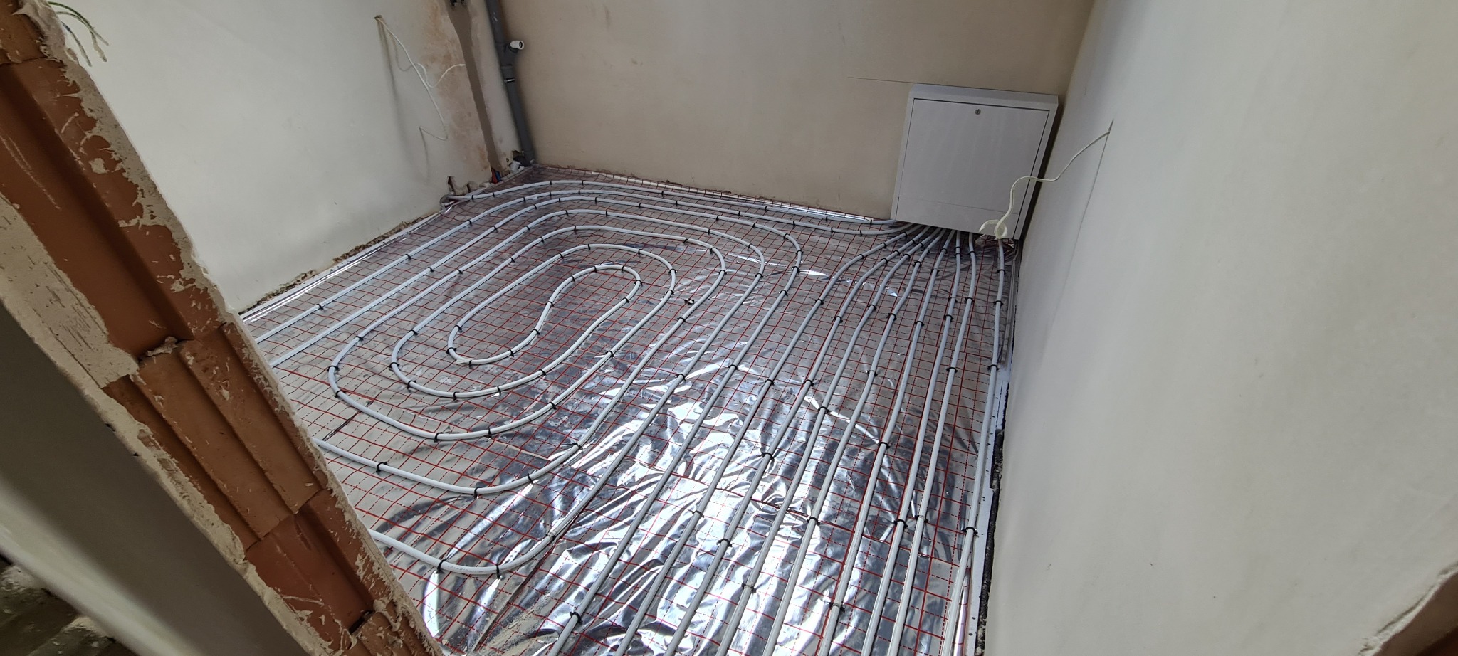 Białe rury ogrzewania podłogowego ułożone na srebrnej folii z czerwoną siatką w surowym pomieszczeniu przed wylewką, widoczna instalacja elektryczna i fragment ceglanego nadproża.