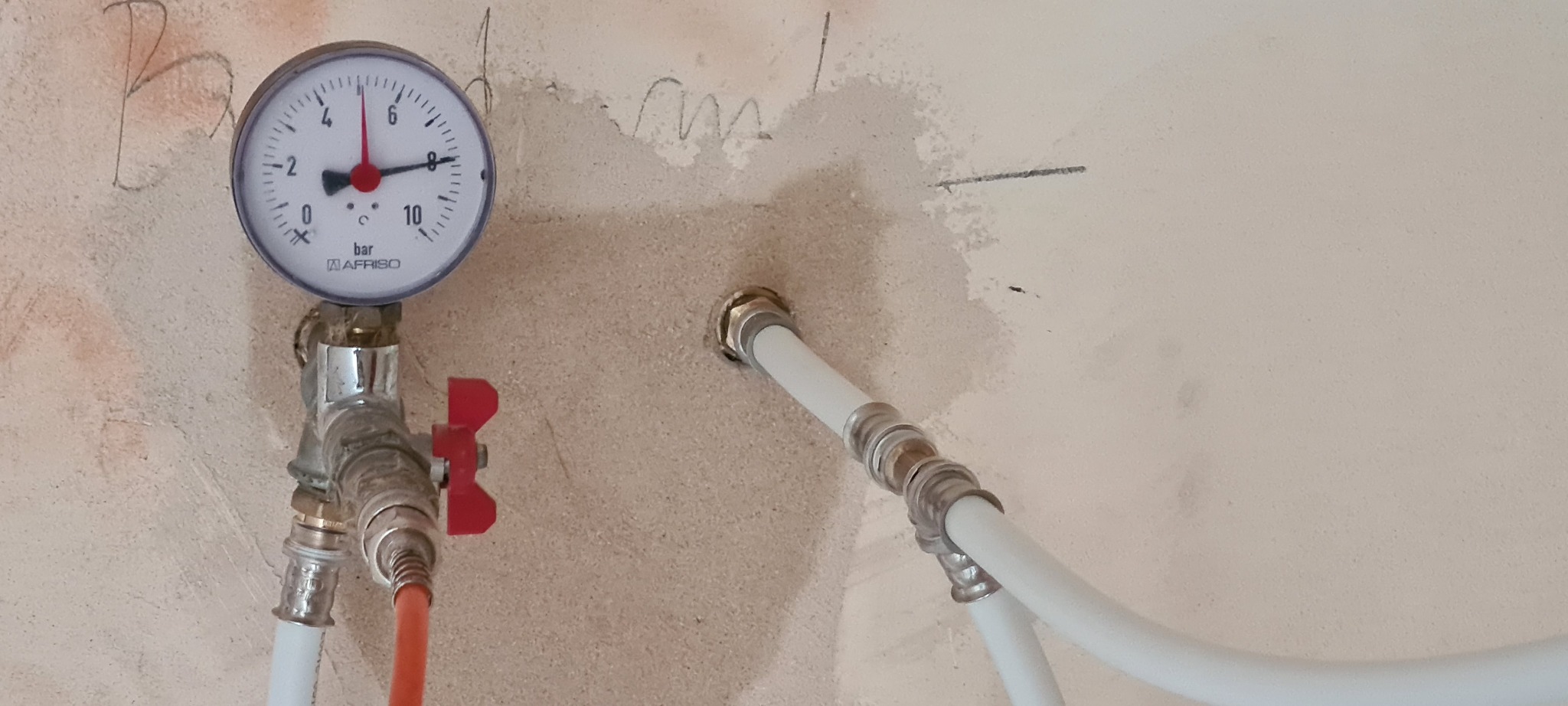 Manometr Afriso zamontowany na instalacji z czerwonym zaworem, widoczne białe rury instalacyjne w surowej ścianie z widocznymi śladami po montażu.