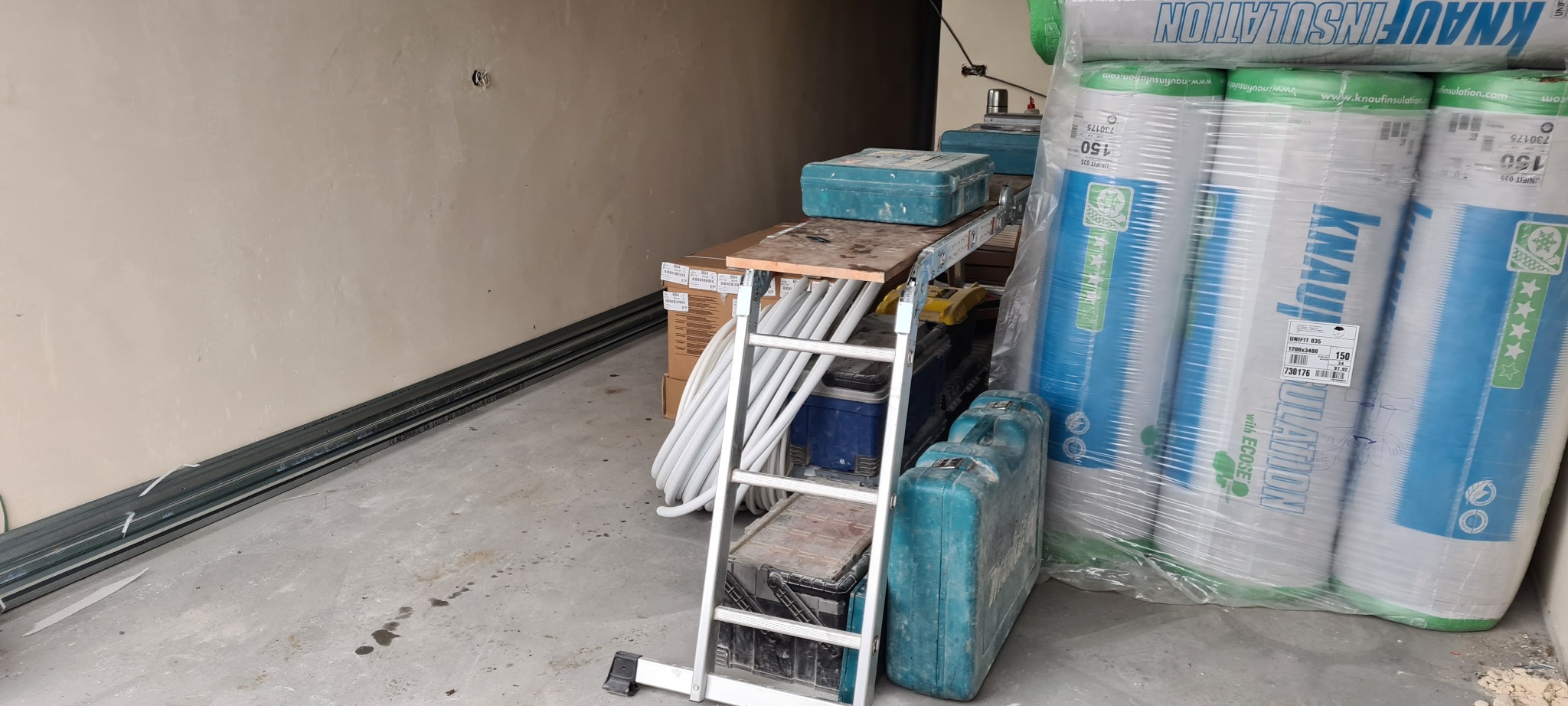 Stos rolek izolacji Knauf, drabina, skrzynki narzędziowe i rury instalacyjne w niezagospodarowanym pomieszczeniu, prawdopodobnie podczas prac remontowych.