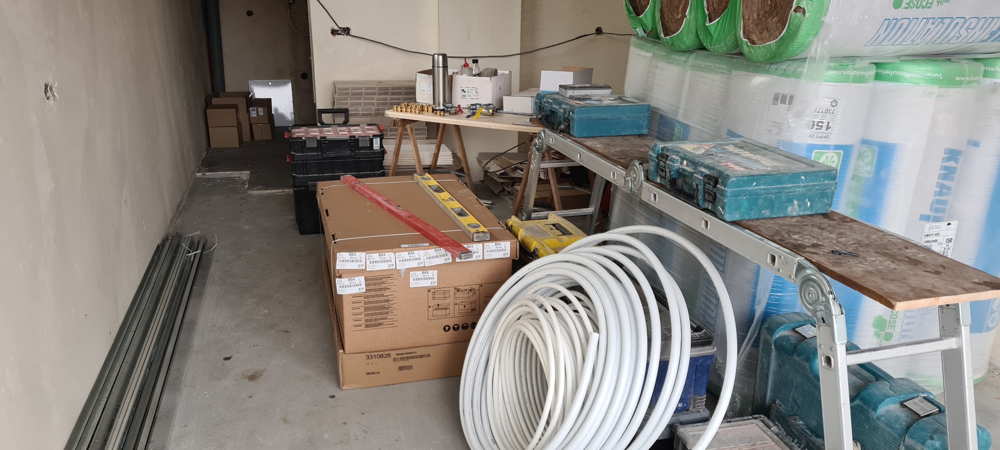 Przygotowania do prac instalacyjnych: zwój białych rur, skrzynki z narzędziami Makita, poziomice na kartonach, izolacja termiczna Knauf na tle surowego wnętrza.