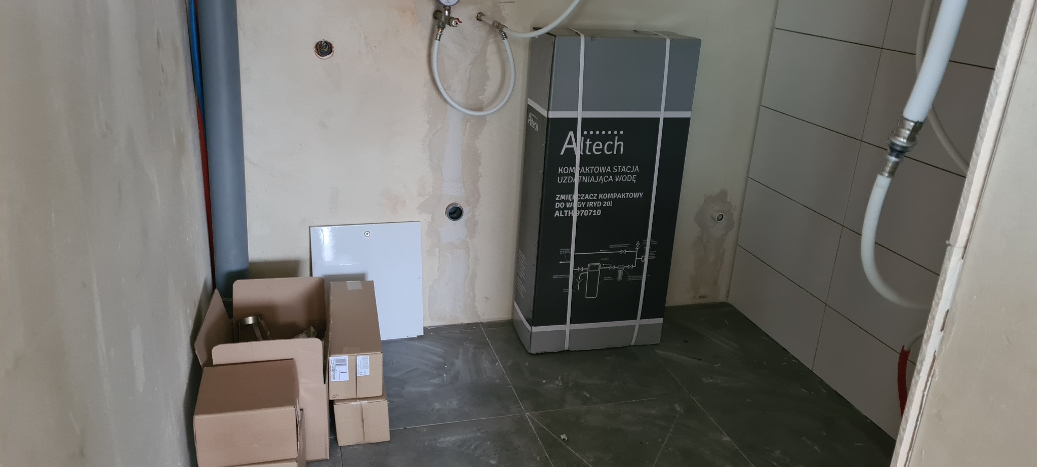 Kompaktowa stacja uzdatniania wody Altech w kartonie, przygotowana do montażu w pomieszczeniu z surowymi ścianami i częściowo wykończoną glazurą.