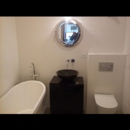 Mastora - Biała łazienka z wanną, umywalką w kształcie misy na czarnej szafce, okrągłym lustrem w stylu bulaja i podwieszaną toaletą.