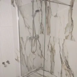 Nowoczesna kabina prysznicowa z chromowaną armaturą i szklaną ścianką, wykończona płytkami imitującymi marmur z delikatnymi żyłami, w minimalistycznej łazience z dekoracyjną ścianą z pionowymi...