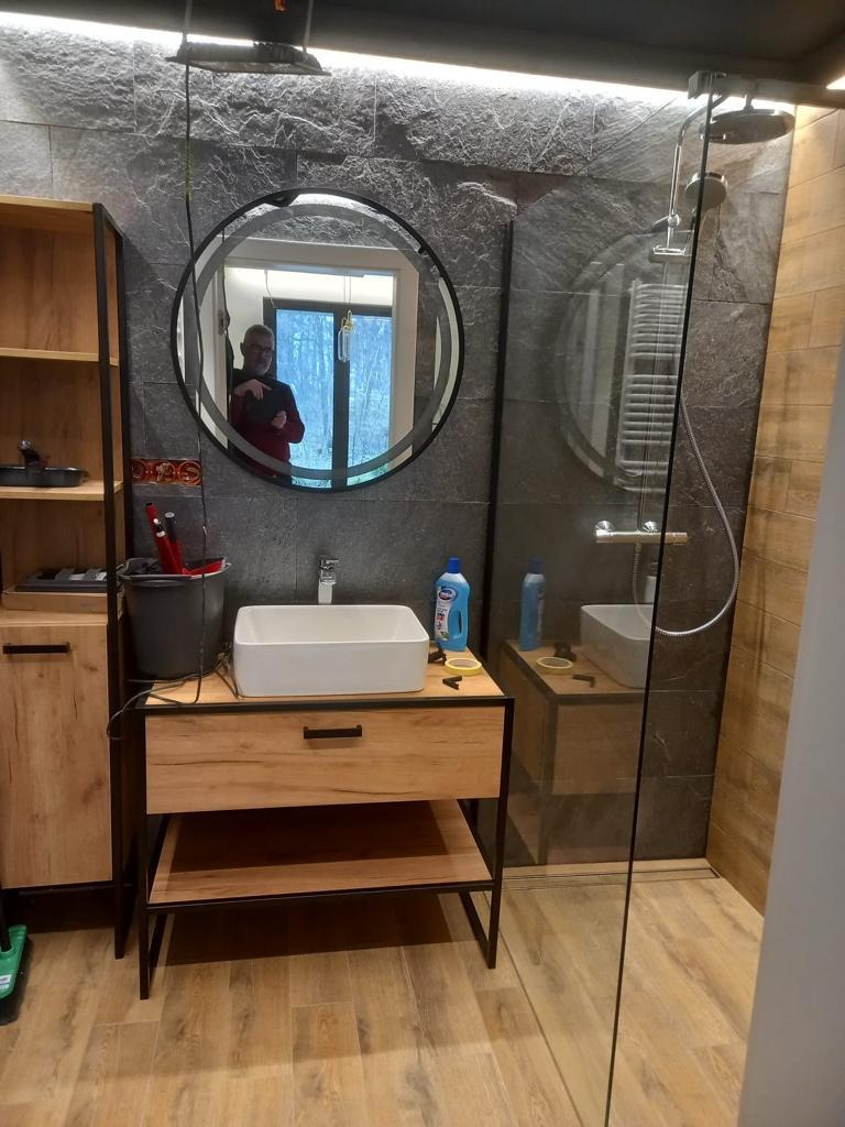 Nowoczesna łazienka z kabiną prysznicową typu walk-in, umywalką nablatową na szafce z drewnopodobnym frontem i okrągłym lustrem z podświetleniem. Na ścianie szara okładzina imitująca kamień.