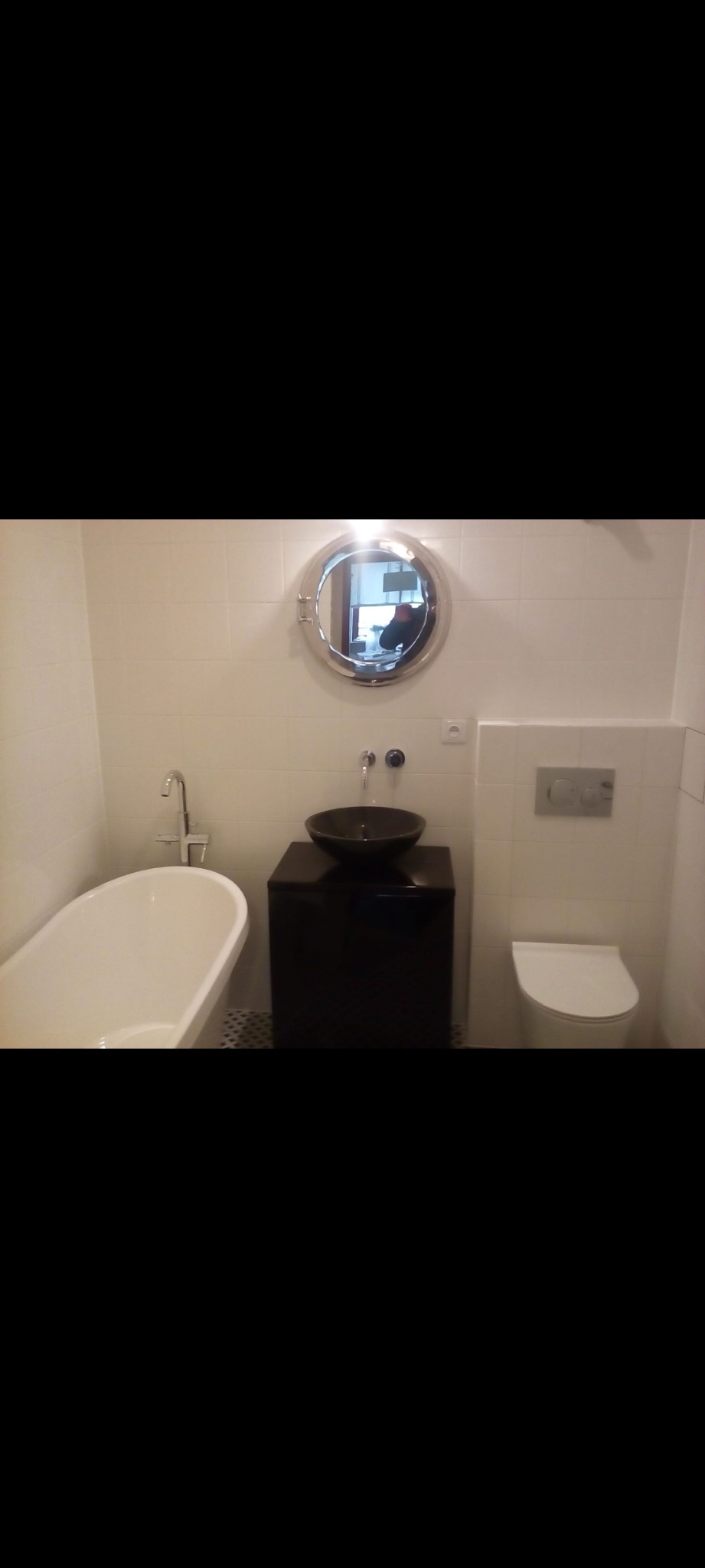 Biała łazienka z wanną, umywalką w kształcie misy na czarnej szafce, okrągłym lustrem w stylu bulaja i podwieszaną toaletą.