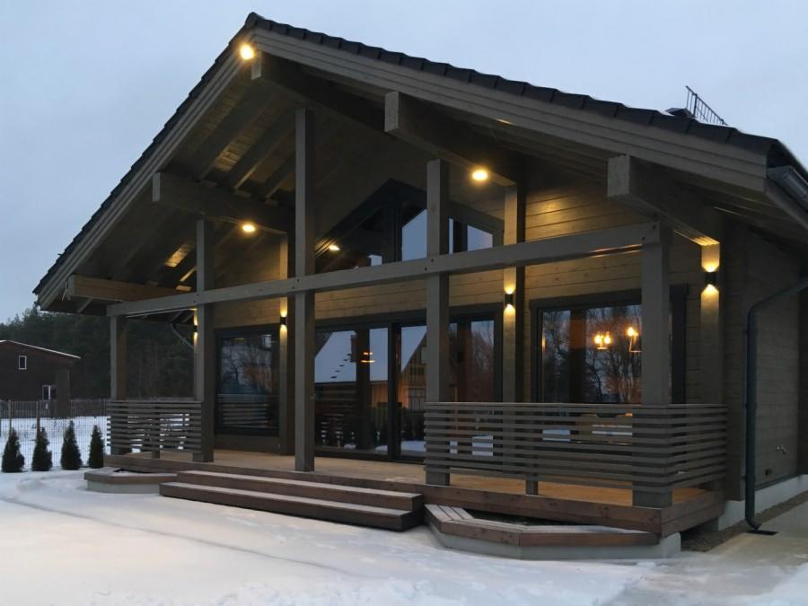 Nowoczesny, drewniany dom z ciemnego drewna w zimowej scenerii, oświetlony zewnętrznymi lampami, z widocznym tarasem i schodami wejściowymi pokrytymi śniegiem.