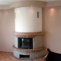 Nowoczesny kominek z obudową z jasnego kamienia i cegły, zwieńczony drewnianą belką, wkomponowany w cylindryczną konstrukcję z wbudowanymi głośnikami, oświetlony punktowo na tle gładkich, beżowych...
