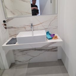 P.M.KOMPLEX - Nowoczesna łazienka z białą umywalką nablatową, marmurowymi płytkami na ścianie i lustrem, w którym odbija się osoba fotografująca wnętrze.