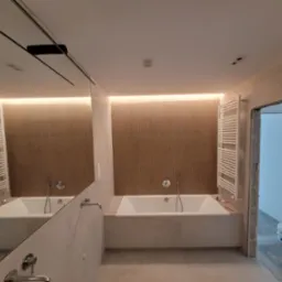 Nowoczesna łazienka z wanną w zabudowie, grzejnikiem drabinkowym i dużym lustrem z czarną ramką, widoczny fragment surowej ściany i wiadro z farbą w sąsiednim pomieszczeniu.