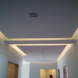 P.M.KOMPLEX - Nowoczesny sufit podwieszany z oświetleniem LED w korytarzu, widoczne fragmenty ścian i otwory drzwiowe w trakcie remontu.