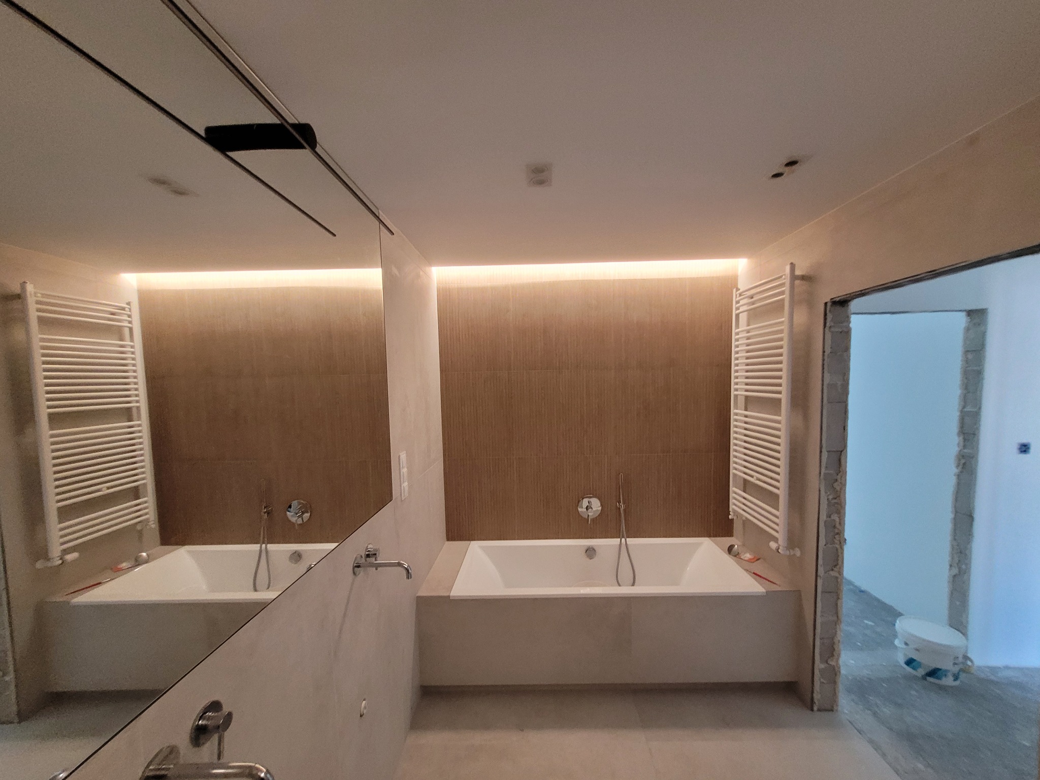 Nowoczesna łazienka z wanną w zabudowie, grzejnikiem drabinkowym i dużym lustrem z czarną ramką, widoczny fragment surowej ściany i wiadro z farbą w sąsiednim pomieszczeniu.