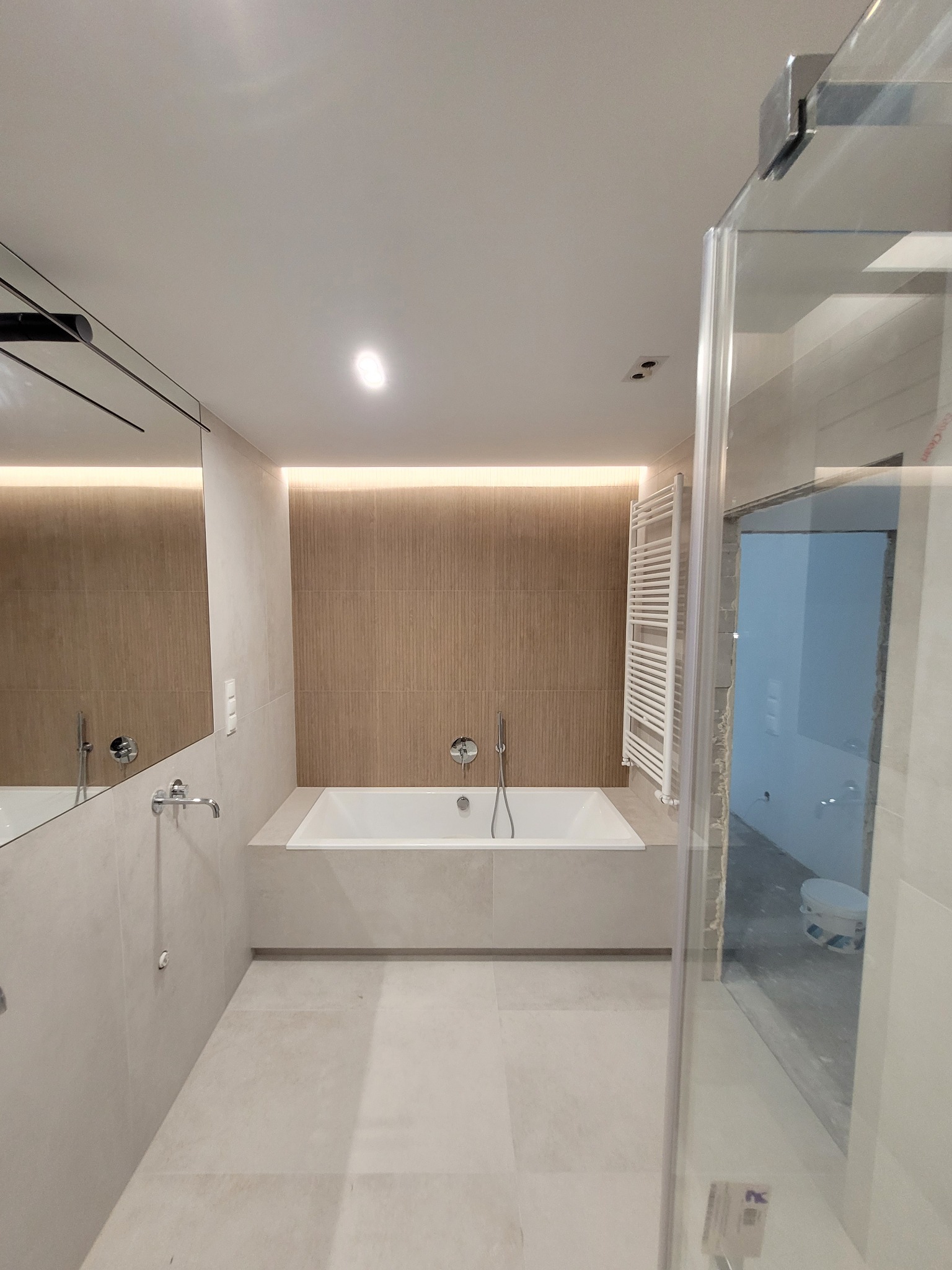 Nowoczesna łazienka z prostokątną wanną w zabudowie, beżowe płytki na ścianach i podłodze, grzejnik drabinkowy oraz częściowo widoczna szklana kabina prysznicowa w trakcie montażu.