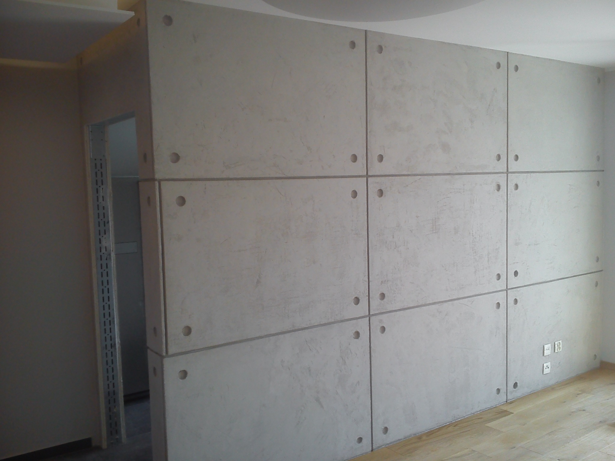 Ściana wykończona płytami betonowymi z otworami, widoczne gniazdka elektryczne i fragment drewnianej podłogi oraz otwarte przejście do innego pomieszczenia z metalowym regałem.