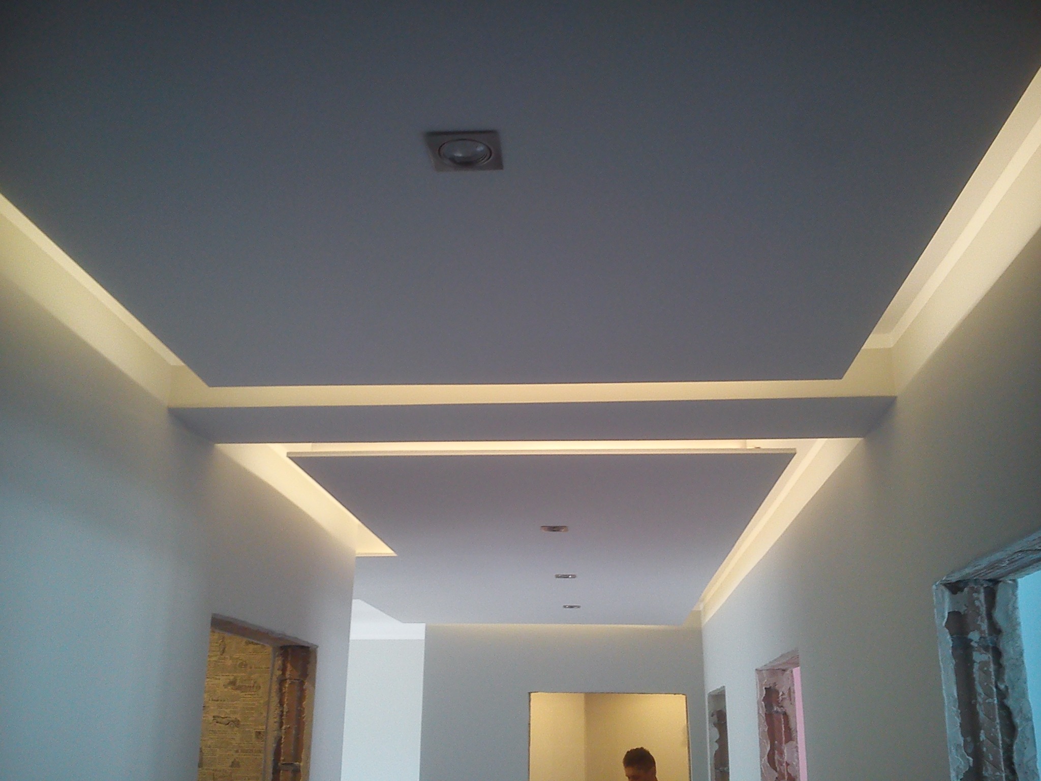 Nowoczesny sufit podwieszany z oświetleniem LED w korytarzu, widoczne fragmenty ścian i otwory drzwiowe w trakcie remontu.