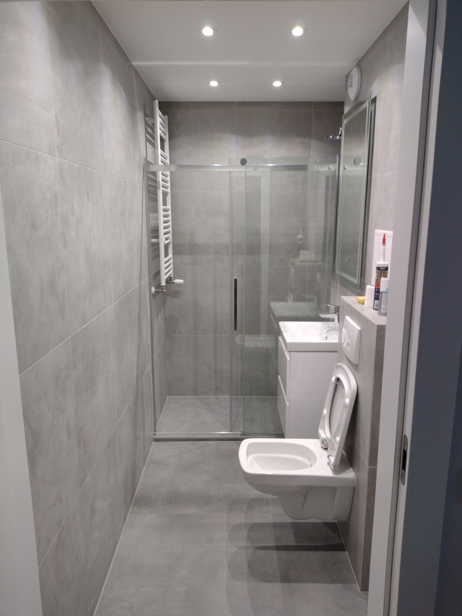 Nowoczesna łazienka w odcieniach szarości z kabiną prysznicową ze szklanymi drzwiami przesuwnymi, grzejnikiem drabinkowym i podwieszaną toaletą z otwartą deską. Na półce nad toaletą widoczne tuby...