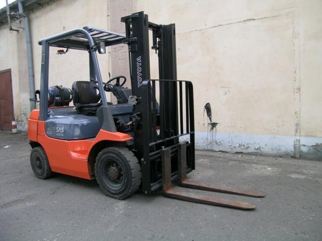 Używany wózek widłowy Toyota 7FGF 25 z 2006 roku, zasilany gazem, stoi na zewnątrz na tle ściany z widocznymi śladami zabrudzeń i odpryskami farby.