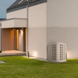 Elewacja nowoczesnego domu z zainstalowaną pompą ciepła, oświetlony ogród, widoczne logo firmy na grafice po prawej stronie.