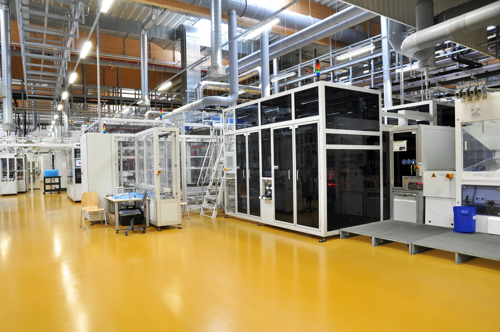 Jasnożółta, bezspoinowa podłoga w hali produkcyjnej z nowoczesnym parkiem maszynowym; widoczne rury wentylacyjne pod sufitem.