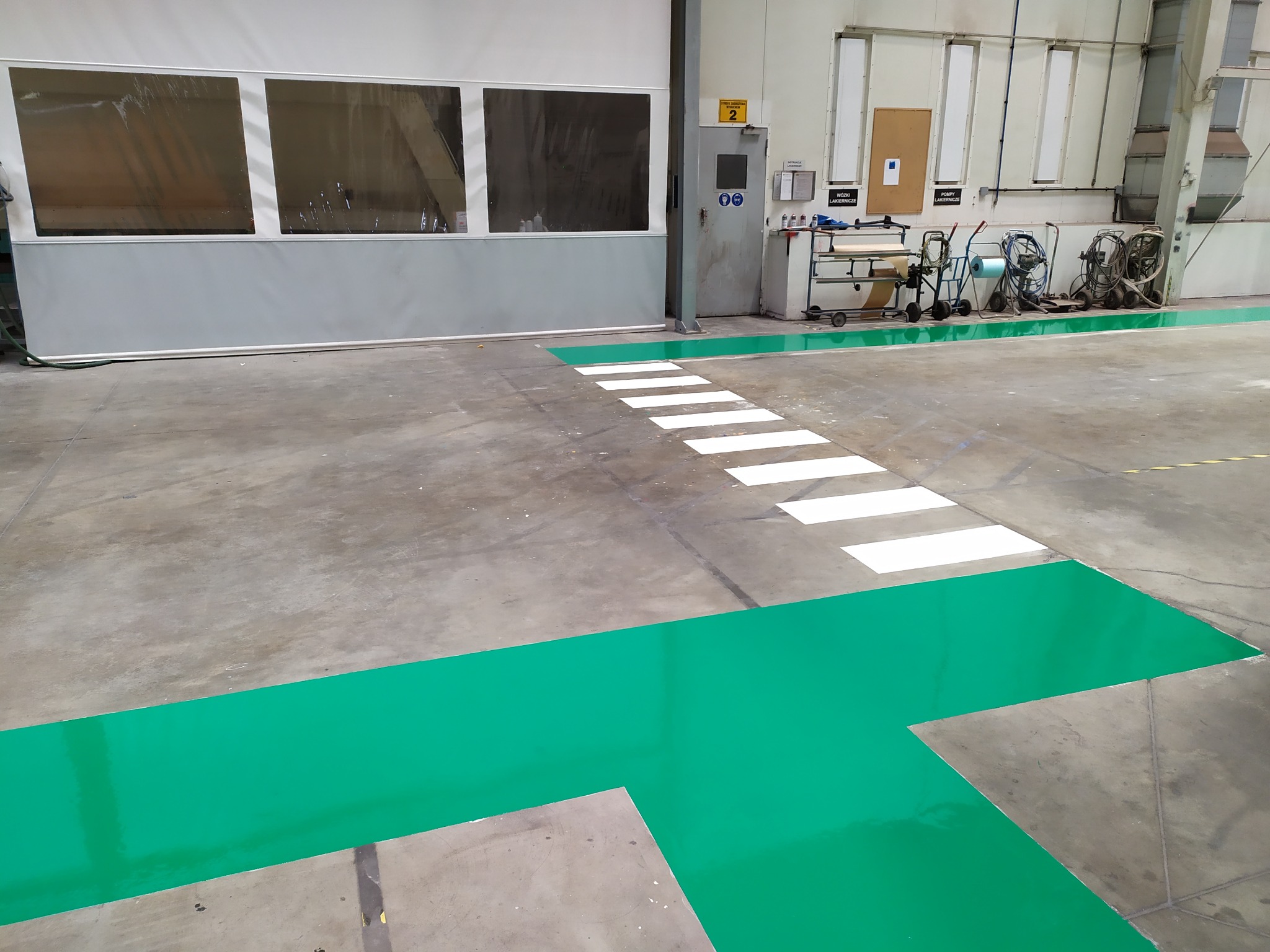 Oznakowanie poziome w hali produkcyjnej: zielone pasy wyznaczające strefy i białe pasy przejścia dla pieszych na szarej posadzce.