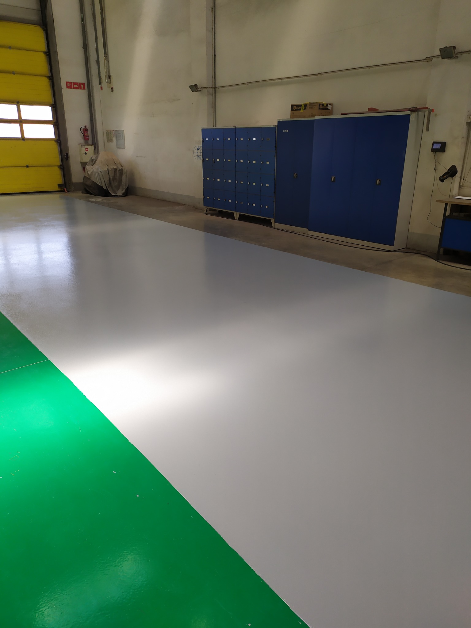 Jasnoszara posadzka w hali produkcyjnej z zielonym fragmentem przy ścianie, widoczne szafki pracownicze i brama wjazdowa.