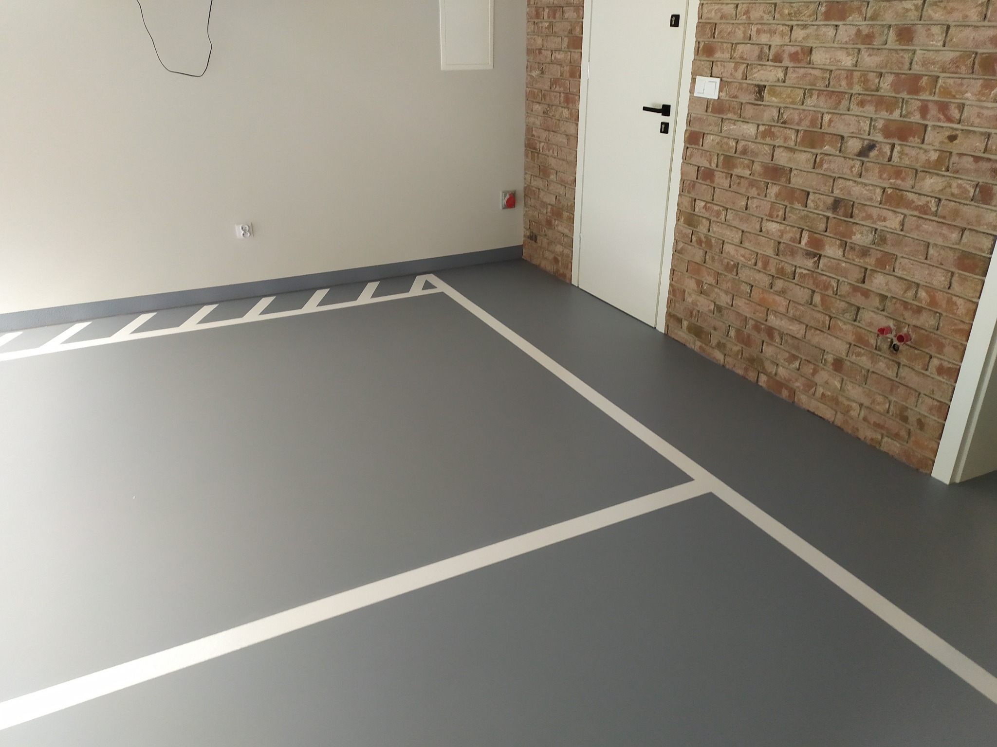 Szara posadzka żywiczna z białymi liniami wyznaczającymi strefy, ściana z cegły i białe drzwi w tle.