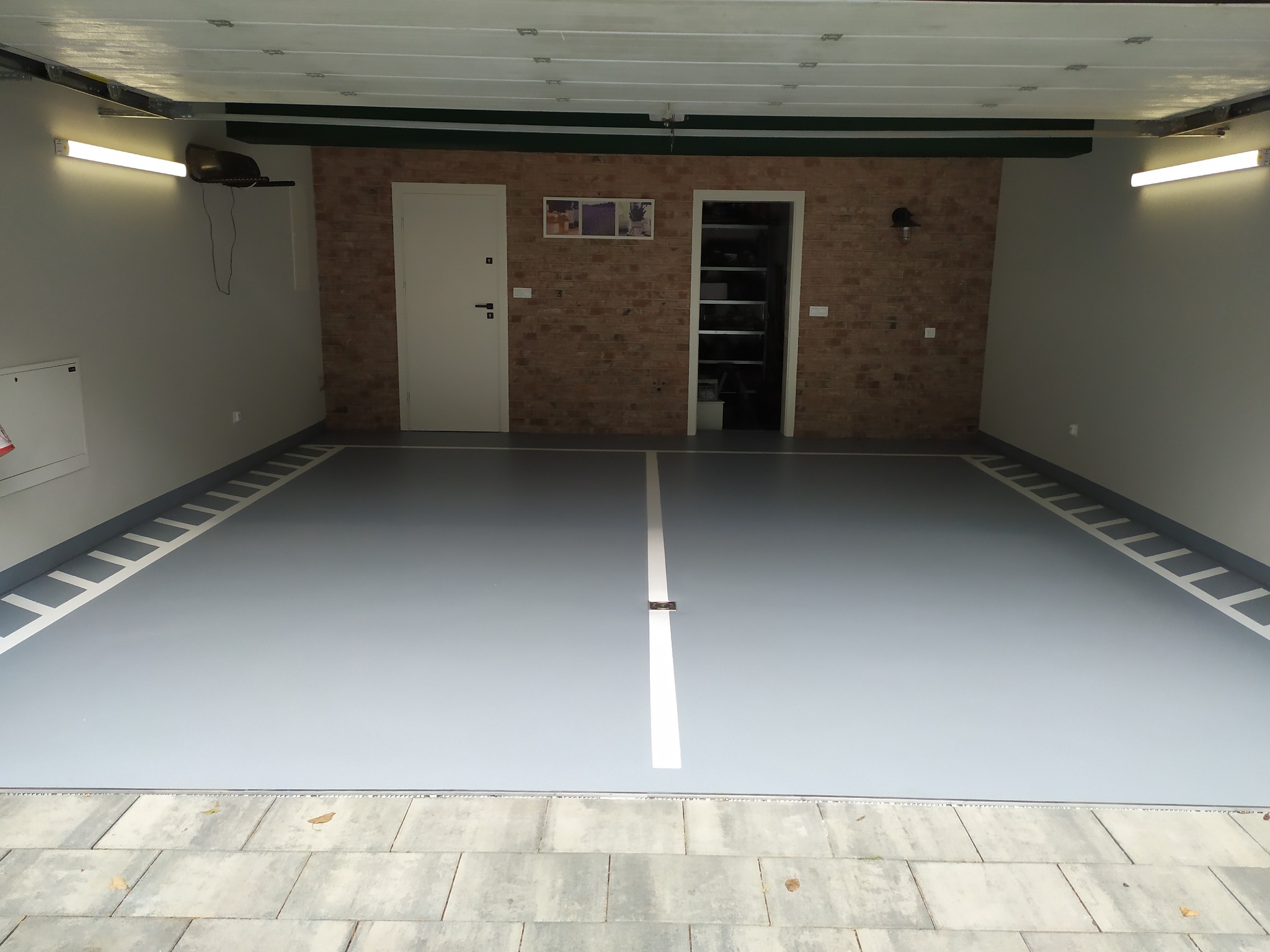 Wnętrze garażu z szarą posadzką, wykończoną białymi liniami i wzorami przypominającymi schody, ceglana ściana z białymi drzwiami i otwartym pomieszczeniem z półkami w tle.