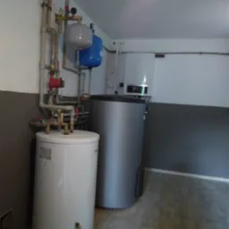 Bojler elektryczny i zasobnik ciepłej wody w kotłowni, widoczne rury instalacji, manometry i zawory.