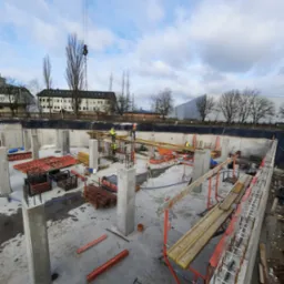 Budowa fundamentów budynku: widoczne betonowe słupy, szalunki, stalowe zbrojenia w ścianach, pracownicy w odblaskowych kamizelkach i dźwig w tle.