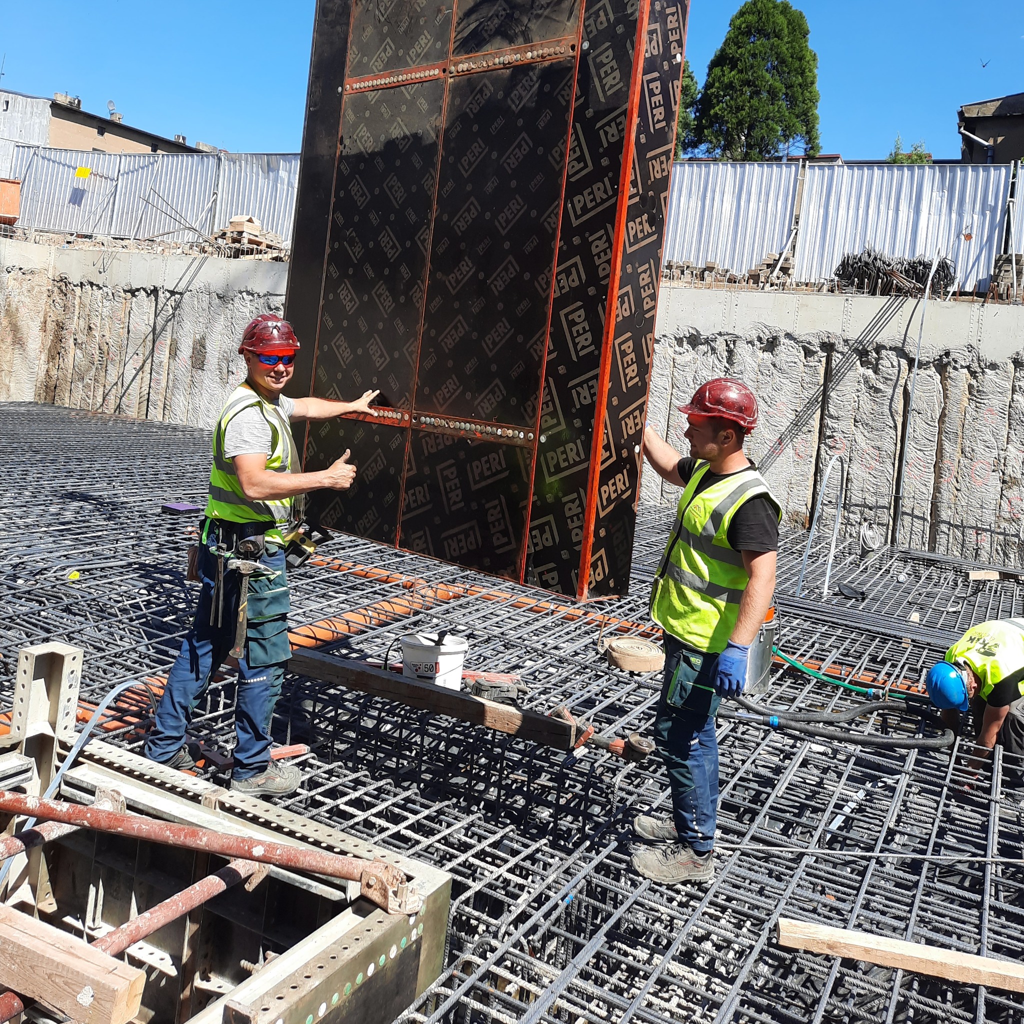 Dwóch pracowników budowlanych w kaskach i kamizelkach odblaskowych przenosi duży panel szalunkowy z logo PERI na budowie, widoczne zbrojenie i wykop fundamentowy.