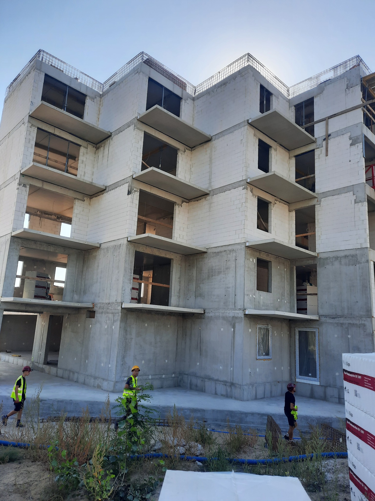 Surowy stan wielopiętrowego budynku z betonu komórkowego, widoczne zbrojenia na dachu, pracownicy w odblaskowych kamizelkach na placu budowy, palety materiałów budowlanych w tle.