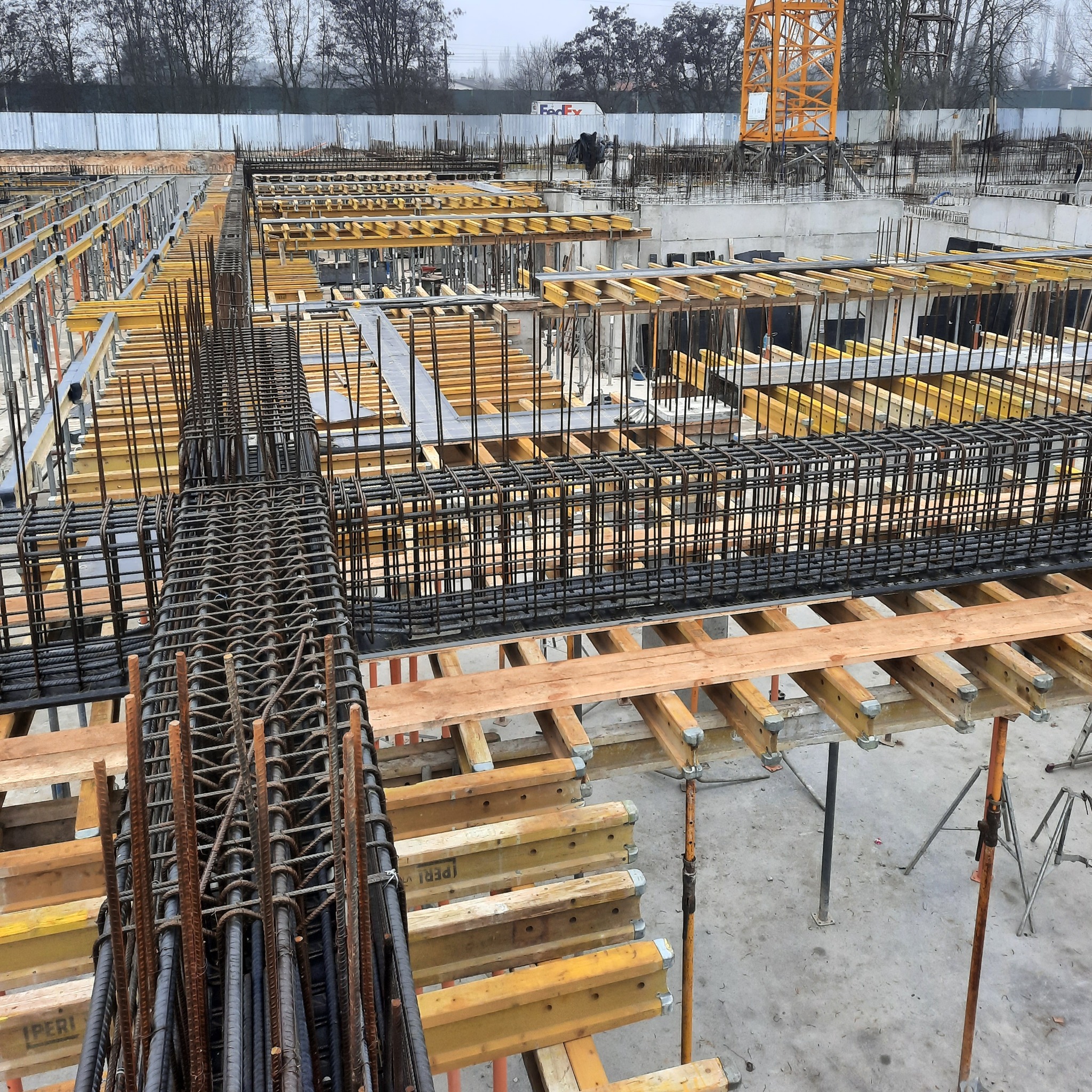 Siatka z prętów zbrojeniowych ułożona na drewnianych podporach na placu budowy, w tle widoczny dźwig i budynek z logo FedEx.