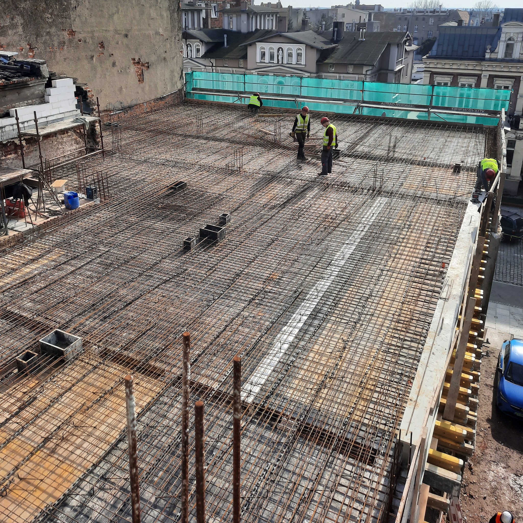 Siatka zbrojeniowa na budowie z pracownikami w kamizelkach odblaskowych, widok z góry na przygotowywaną konstrukcję stropu, w tle zabudowa miejska.