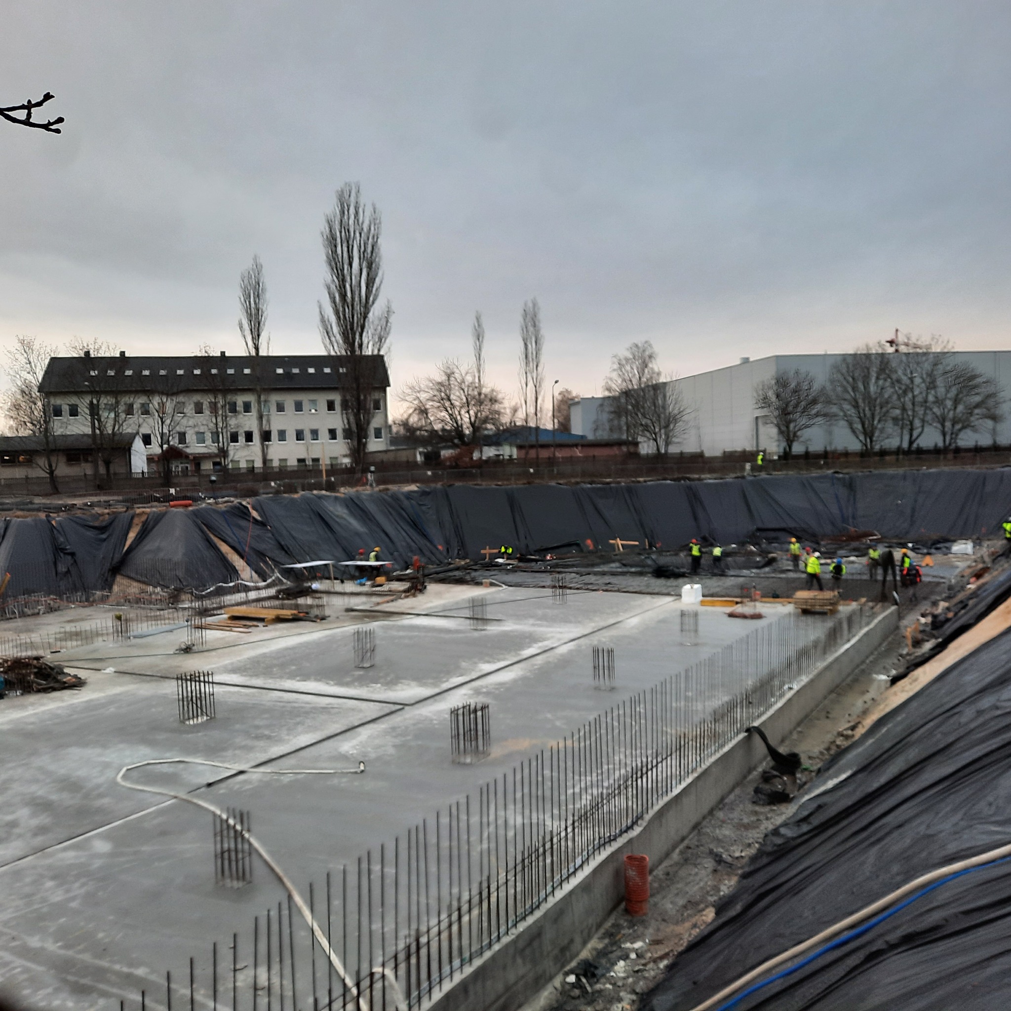 Wylana betonowa płyta fundamentowa z wystającymi prętami zbrojeniowymi, zabezpieczona czarną folią, budynek w tle, pochmurny dzień.