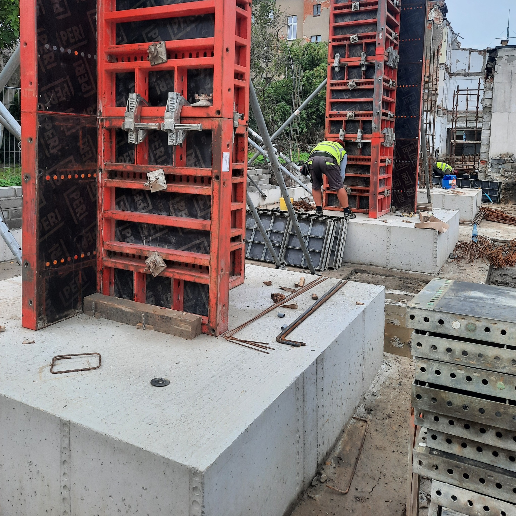 Montaż szalunków z czerwonymi ramami na betonowych fundamentach, pracownik w kamizelce odblaskowej w tle, widoczne elementy zbrojeniowe i pozostałości po zburzonym budynku.