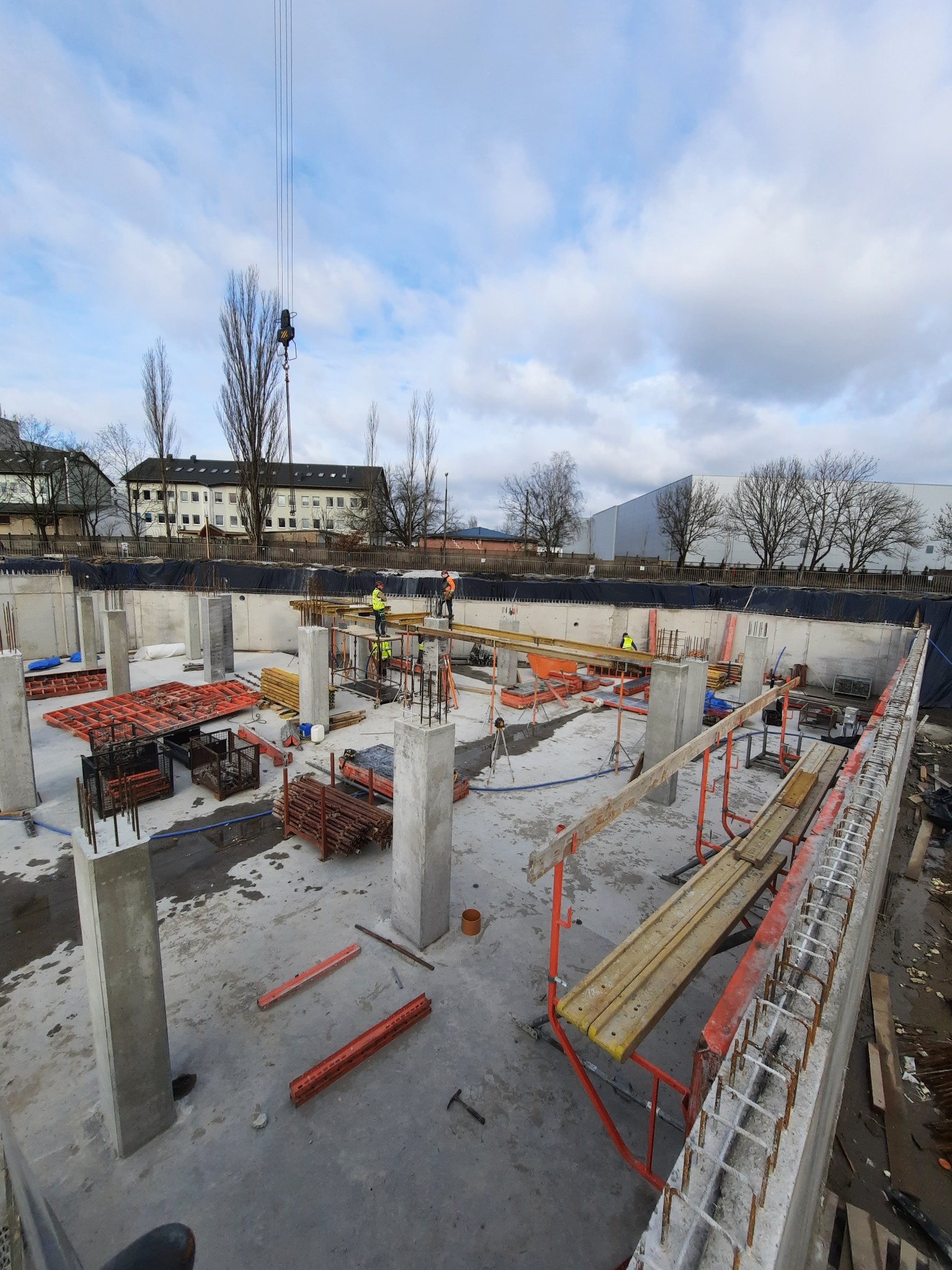 Budowa fundamentów budynku: widoczne betonowe słupy, szalunki, stalowe zbrojenia w ścianach, pracownicy w odblaskowych kamizelkach i dźwig w tle.