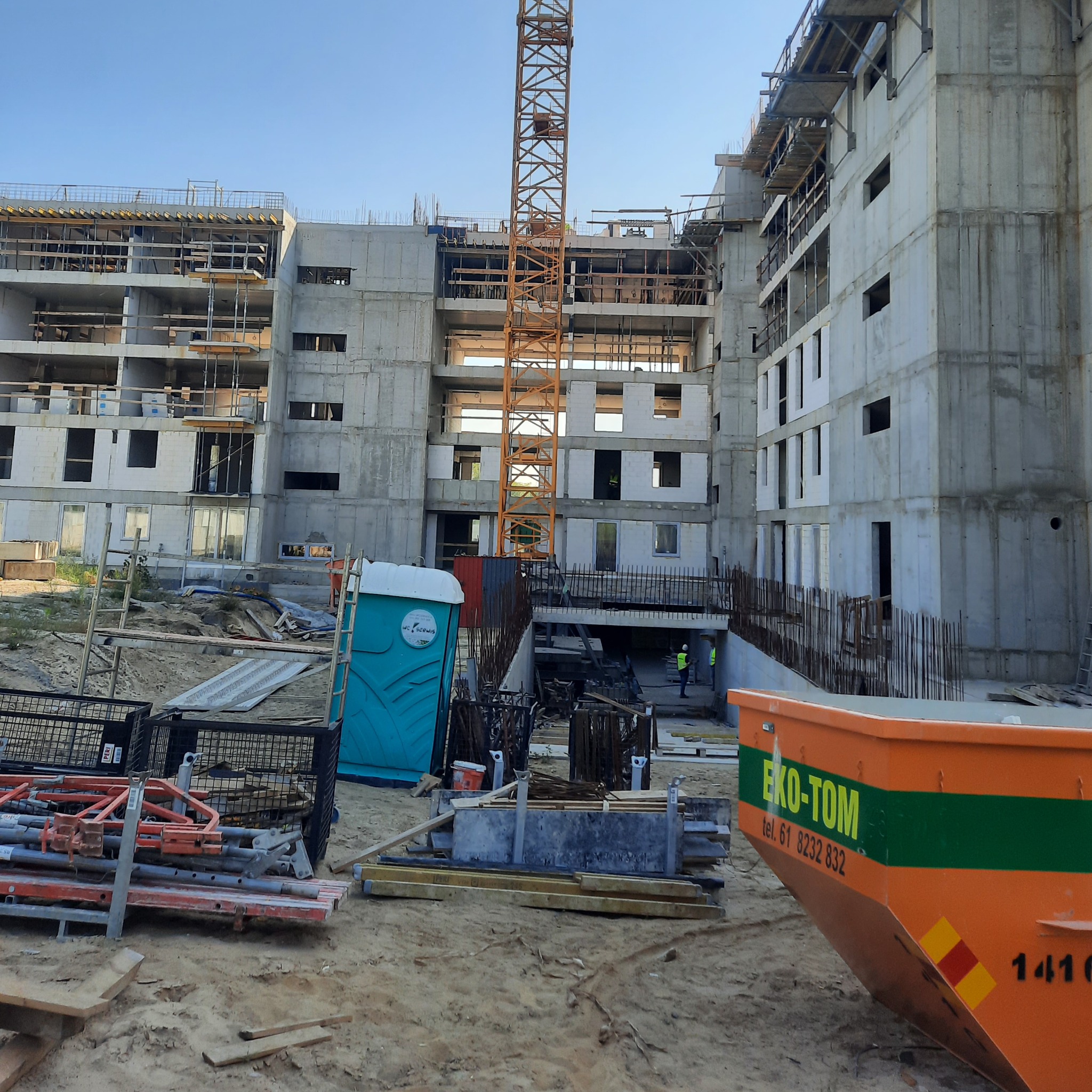Wnętrze placu budowy z widokiem na budynek w surowym stanie, żuraw wieżowy, przenośną toaletę i kontener na gruz z napisem EKO-TOM.