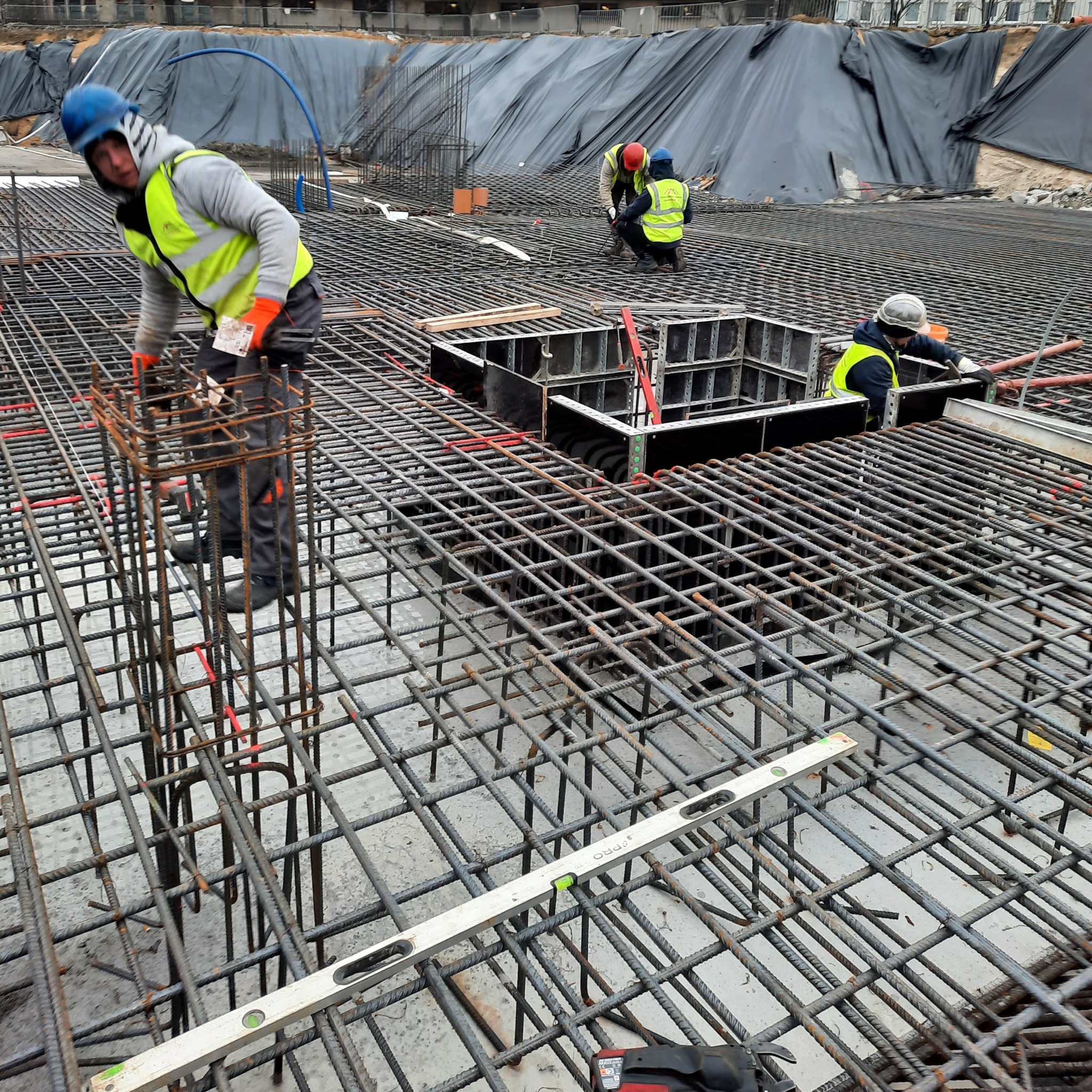 Prace na budowie: kilku pracowników układa siatkę zbrojeniową z prętów stalowych, widoczny poziomica, szalunki i zabezpieczenie wykopu folią.