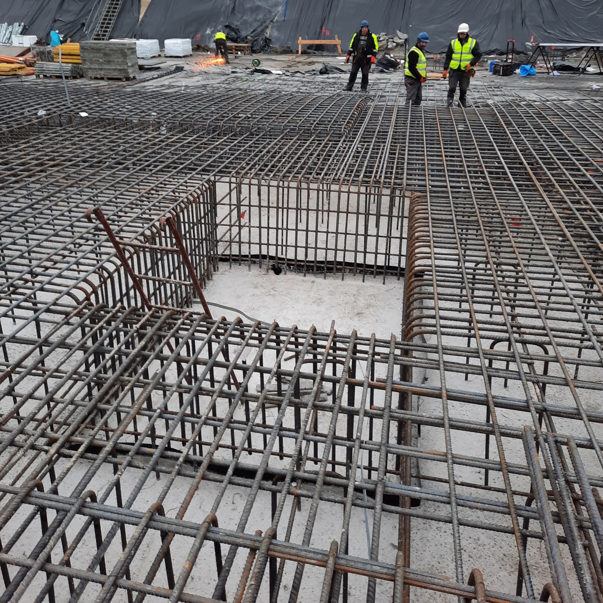Siatka zbrojeniowa na placu budowy z widocznymi pracownikami w oddali, w trakcie spawania prętów, z wycięciem w siatce.