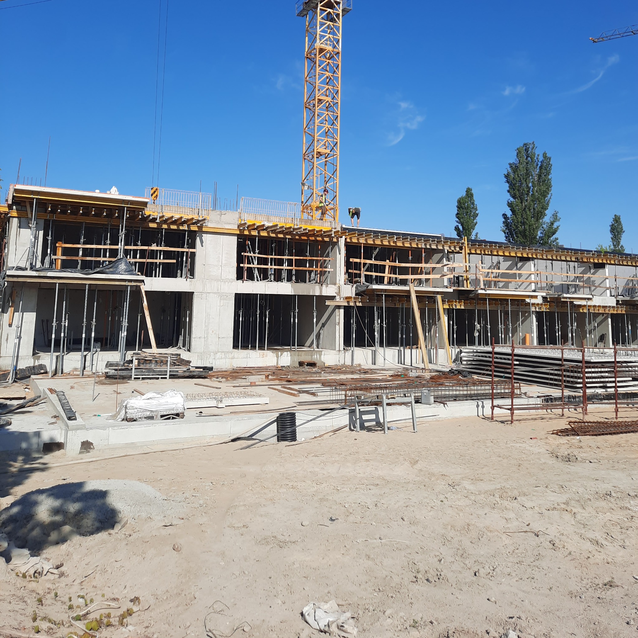 Budowa wielopiętrowego budynku w trakcie wznoszenia konstrukcji żelbetowej, widoczne szalunki, zbrojenia i dźwig na tle błękitnego nieba.