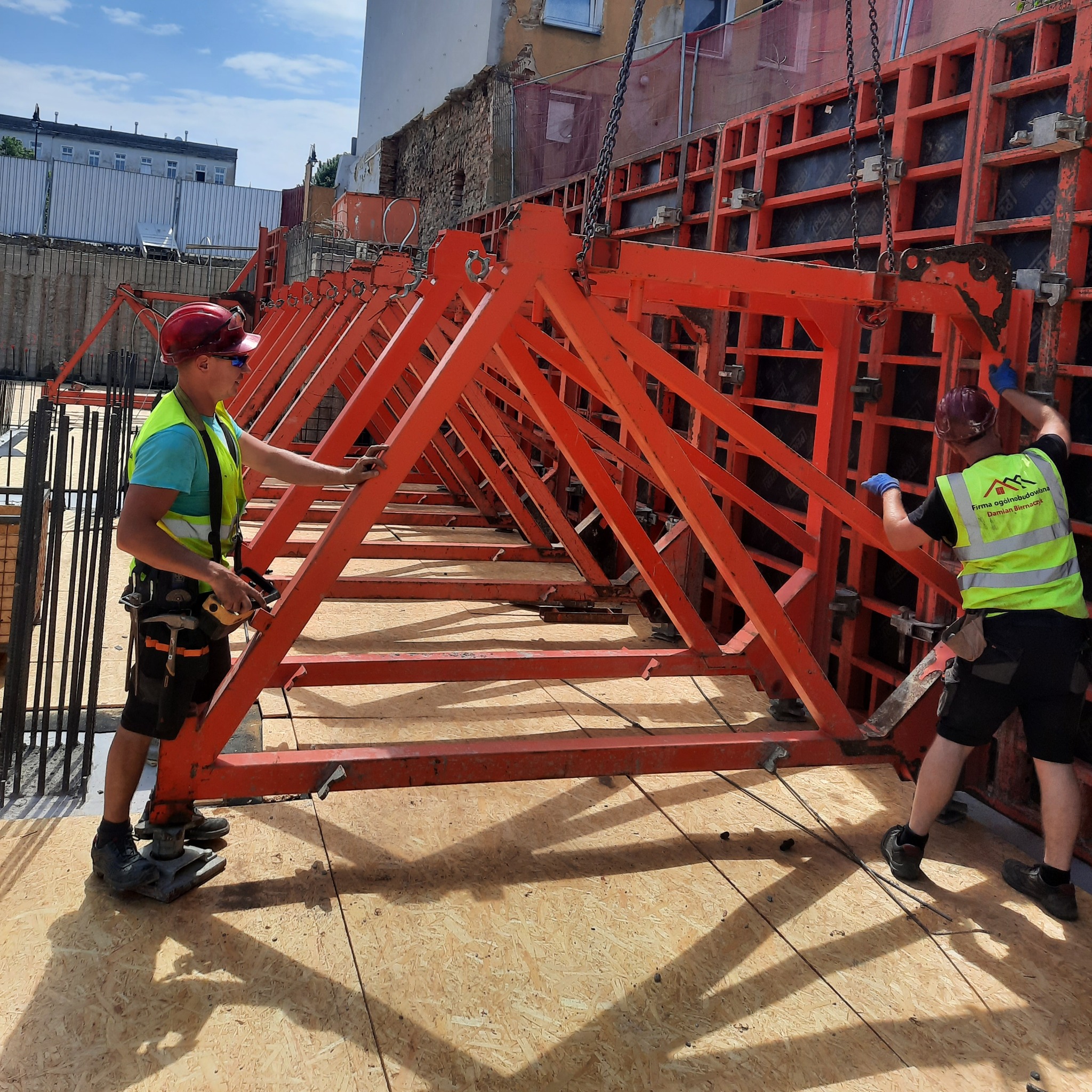 Dwóch zbrojarzy w kaskach i kamizelkach odblaskowych montuje pomarańczowe szalunki na placu budowy, widoczne zbrojenie w tle.