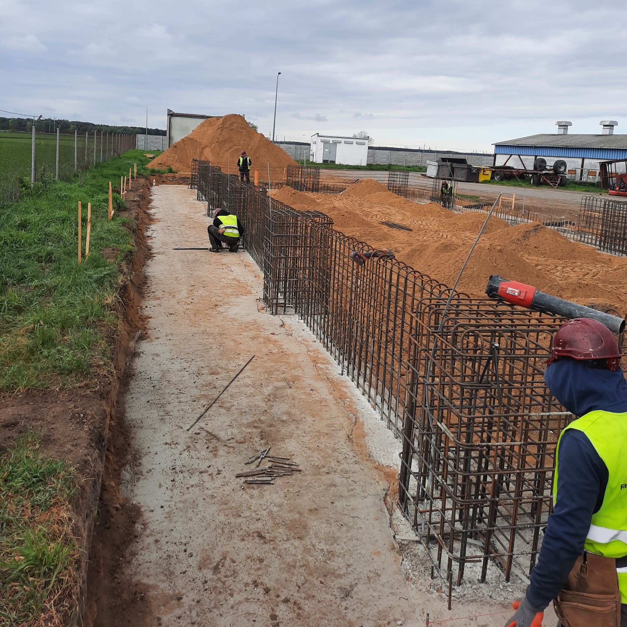 Przygotowanie zbrojenia fundamentów pod budynek przemysłowy, widoczni pracownicy w odblaskowych kamizelkach, narzędzia i hałda piasku w tle.