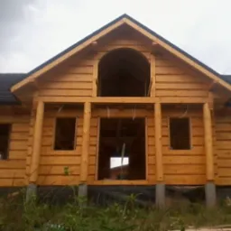 Surowy stan domu z bali z ciemną dachówką i charakterystycznym łukowym oknem na poddaszu, wsparty na betonowych fundamentach w otoczeniu zieleni.