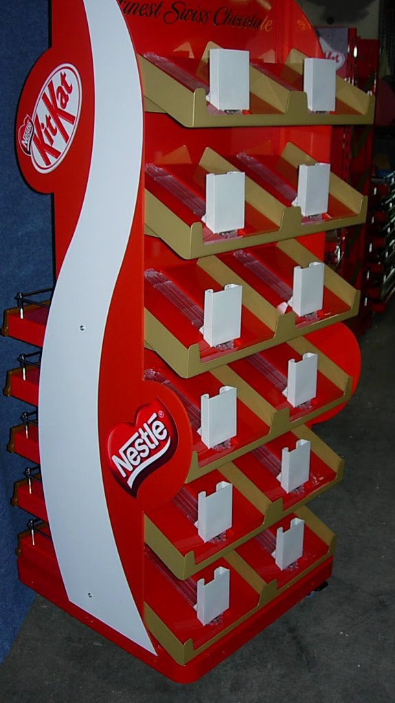 Czerwono-biały stojak reklamowy z logo KitKat i Nestle, przeznaczony do ekspozycji produktów w Pucku, z półkami wykończonymi złotym kolorem i białymi elementami mocującymi.