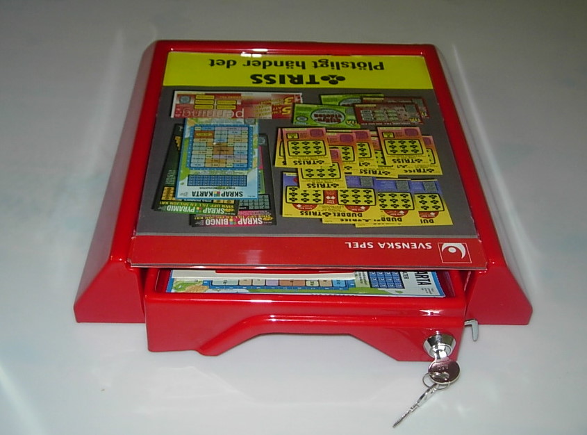 Czerwony ekspozytor na loterie Triss z kluczykiem, prezentujący różne rodzaje zdrapek w górnej części i schowku z zapasem w dolnej szufladzie.