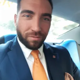 Portret mężczyzny w niebieskim garniturze i pomarańczowym krawacie, siedzącego na niebieskim siedzeniu w pojeździe.