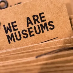 Stos brązowych etykiet z napisem 'WE ARE MUSEUMS' w czcionce bezszeryfowej, z metalowym oczkiem i sznurkiem, ułożonych warstwami.