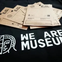 Czarna torba z białym logo 'WE ARE MUSEUMS' i stosem brązowych, papierowych torebek z nadrukiem, leżących na niej. Torebki zawierają informację o wydarzeniu w Muzeum Śląskim w Katowicach w dniach...